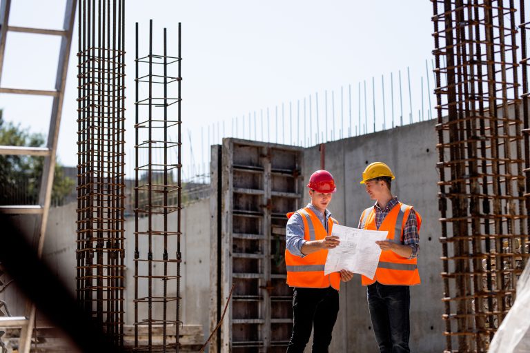 Civil engineer and construction manager in orange work vests and hard helmets explore construction documentation on the building site near the steel frames . .